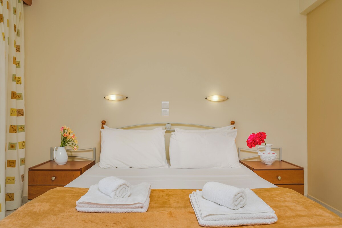 A hotel room with a bed, two nightstands, a night lamp, a vase with flowers, and towels.
