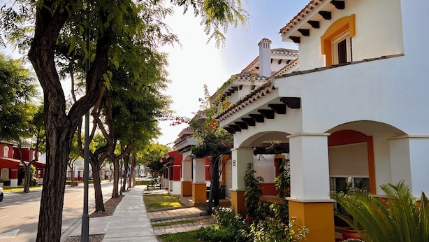 Una hilera de edificios de estilo tradicional con fachadas blancas y amarillas, ventanas arqueadas y una acera bordeada de árboles.