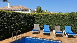 Una piscina con cuatro sillas de descanso, una escalera metálica y un seto al fondo.
