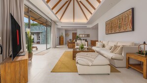 Un salon moderne avec un grand canapé, une table basse et un téléviseur posé sur un meuble en bois. Une grande fenêtre avec des rideaux, un tableau au mur et un plafond en bois complètent la pièce.