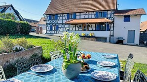 An outdoor dining area with a table set for two, a potted plant, and a view of a traditional half-timbered house. - La Maison «  Schmoutz 💋 »     Elegant Alsatian house dating from 1752 (Forstheim)