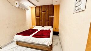 A small, narrow hotel room with a wooden headboard, a bed with white linens and red blankets, a wooden nightstand, and a wall-mounted air conditioning unit. - Hotel A Grand near All Main Places  (Ujjain)