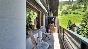 En balkong med trämöbler, bestående av stolar och ett bord, med utsikt över ett grönskande landskap med träd och ett berg.