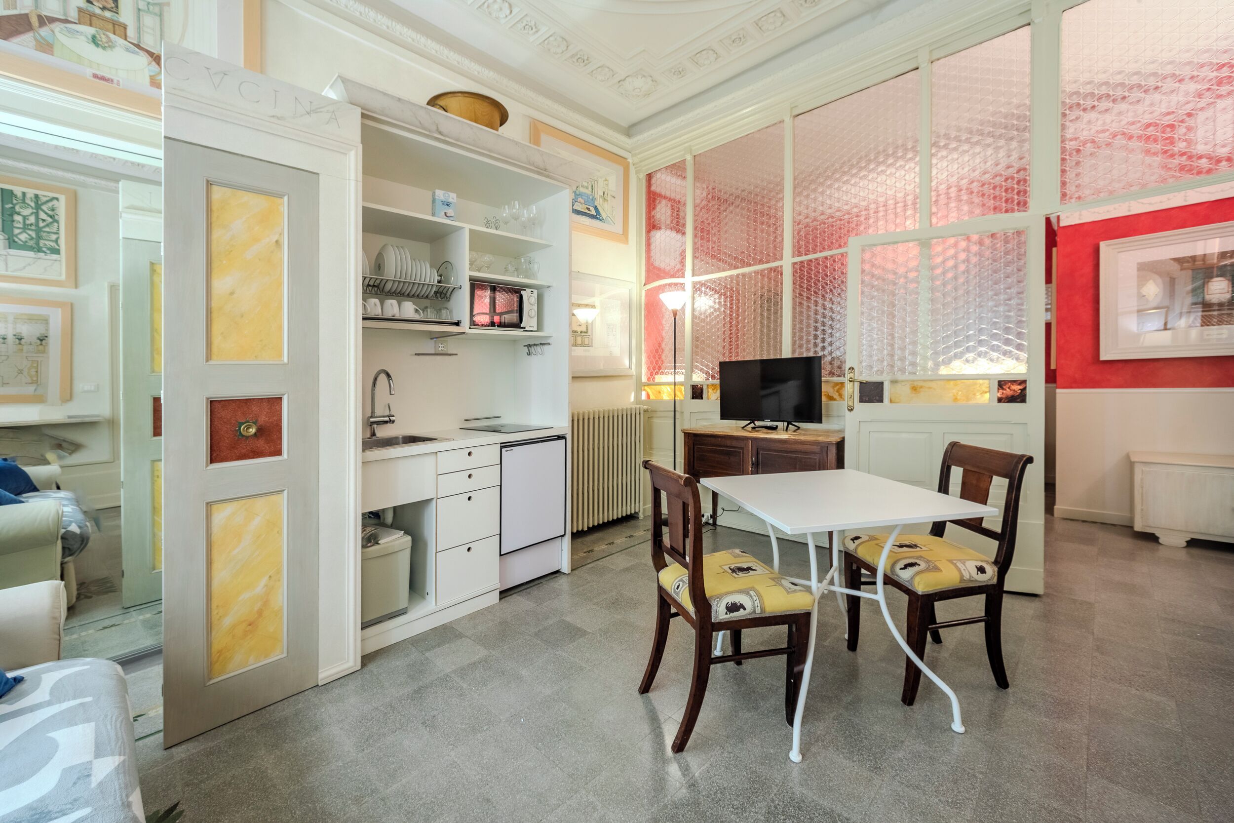 A compact kitchen with a dining area, featuring a white table and chairs, a microwave, and a TV.