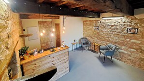 Un intérieur rustique avec des poutres apparentes, un mur en pierre, un comptoir en bois agrémenté d’objets décoratifs et un coin salon avec une chaise et une table.