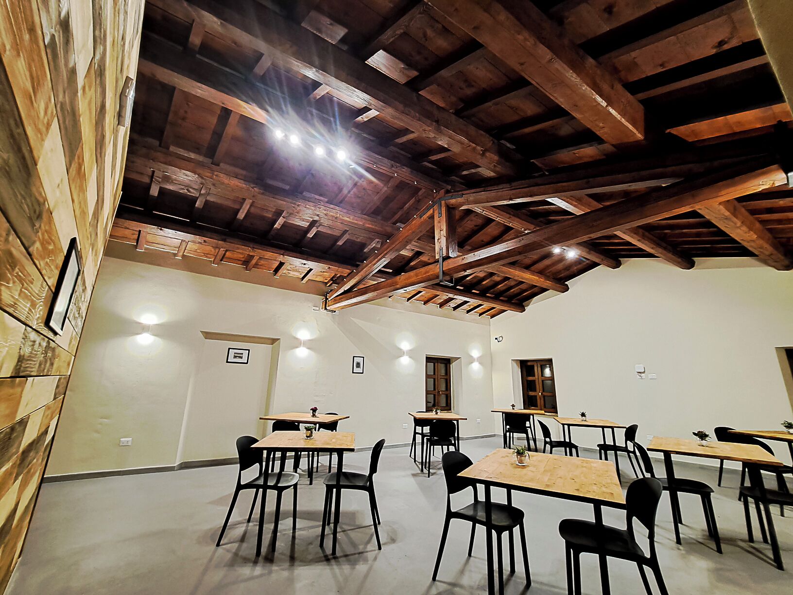 Een ruime kamer met houten balken in het plafond, meerdere tafels met stoelen, en ingelijste foto’s aan de muren.