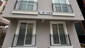A multi-story building with a grey facade, white window frames, and black balcony railings. The name "Call Suit" is displayed on the building.