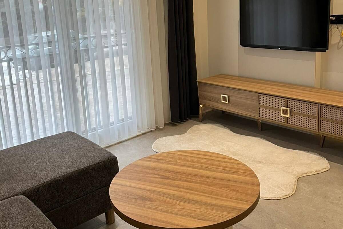 A modern living room with a round wooden coffee table, a dark grey sofa, a wooden TV stand, and a large flat-screen TV mounted on the wall.