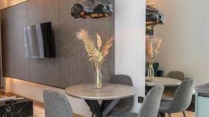 Een moderne eetruimte met een ronde tafel, gestoffeerde grijze stoelen en een minimalistische inrichting, waaronder een vaas met gedroogde planten.
