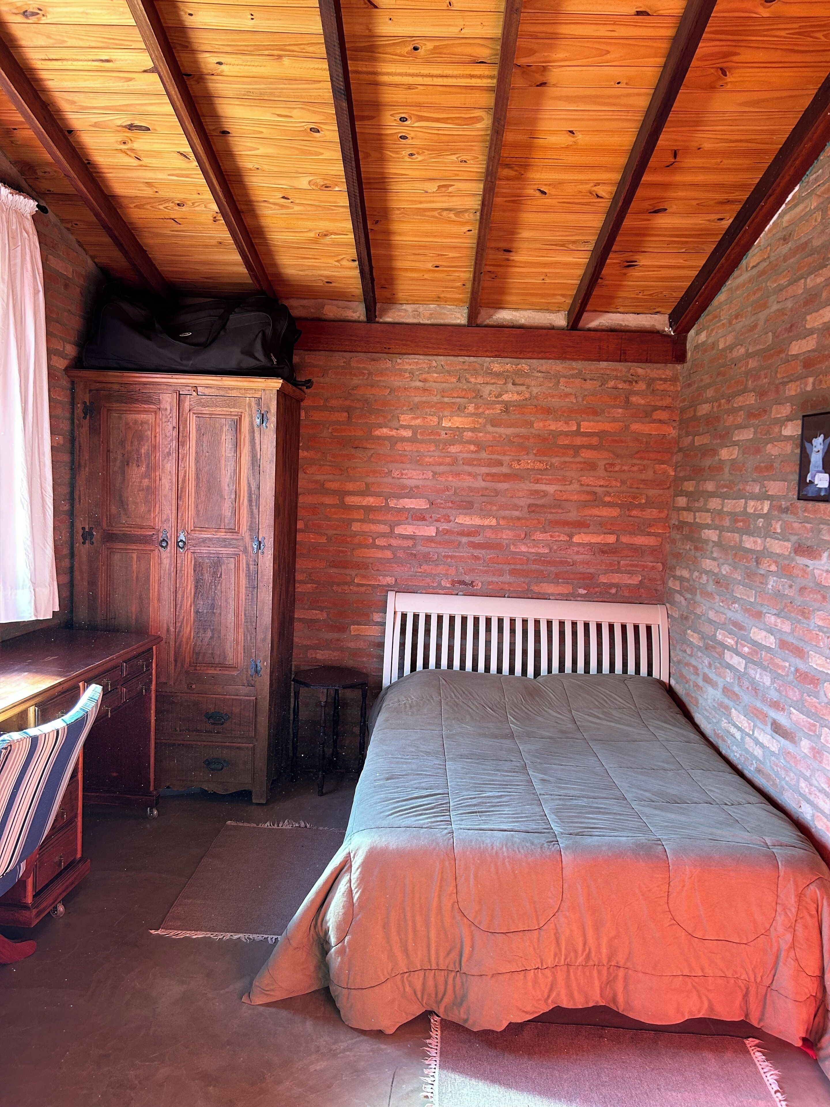 A bedroom with a wooden ceiling, brick walls, a bed with a white headboard, a wooden wardrobe, and a small table.