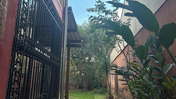 A narrow alleyway with a brick wall on one side and a metal gate. There is a concrete path leading into the alley, surrounded by greenery and a building with a white facade. - Beautiful house in Barão Geraldo (5 min from Unicamp) (Campinas)