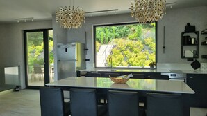 A modern kitchen with a large island, two hanging light fixtures, a refrigerator, and a view of greenery outside.