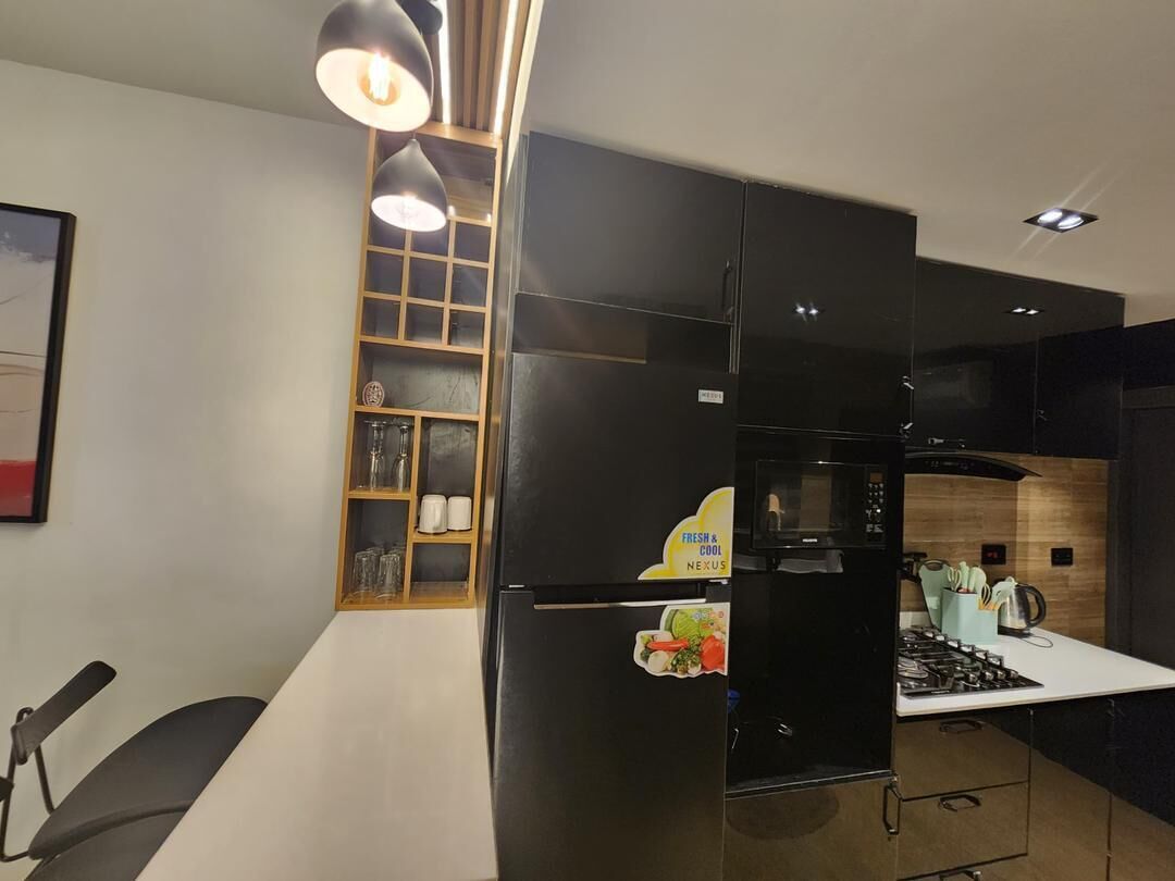 A modern kitchen with a black refrigerator, a microwave, and a stove. There is a wooden shelf unit with glassware and a framed picture on the wall.