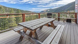 Um deck de madeira com mesa de piquenique e churrasqueira, com vista para uma paisagem montanhosa.