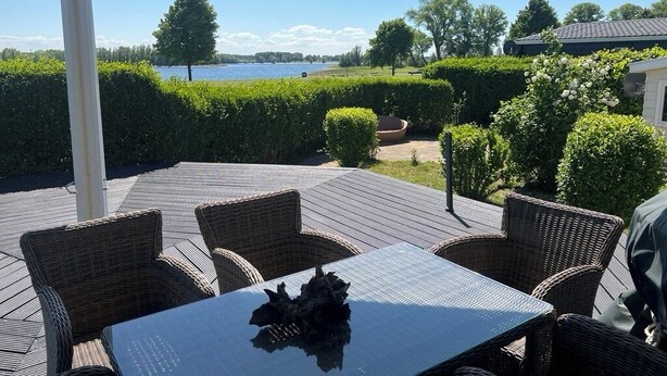 An outdoor patio with wicker furniture and a glass table, overlooking a lake and neatly trimmed hedges.