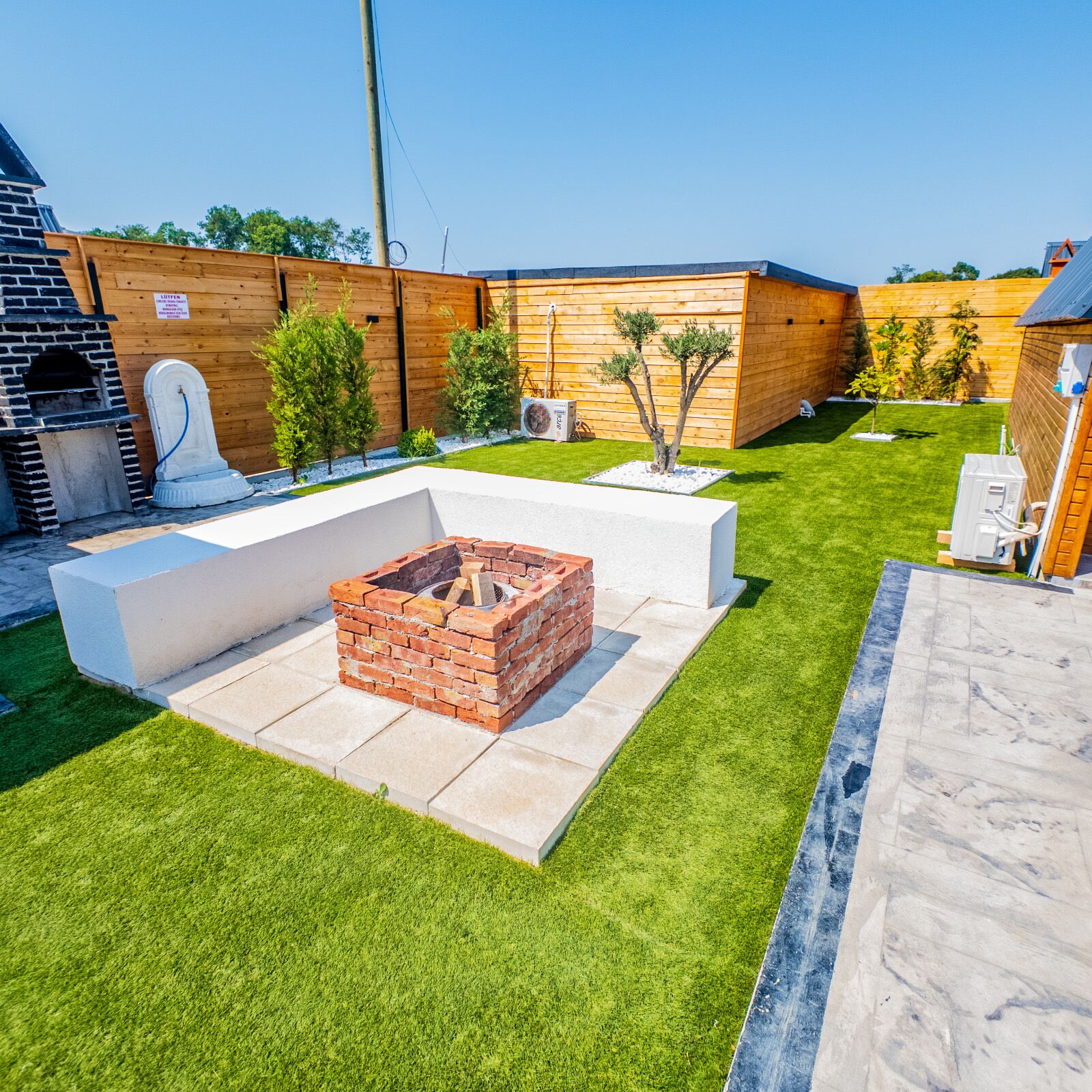 Un jardin avec un foyer en briques, une terrasse en bois et une pelouse verdoyante.
