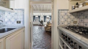 Een keuken met witte kastjes, een spoelbak en een kookplaat. Er is een betegelde muur met een patroon en een visgraatvloer. Een doorgang leidt naar een woonruimte met een bank en een tafel.