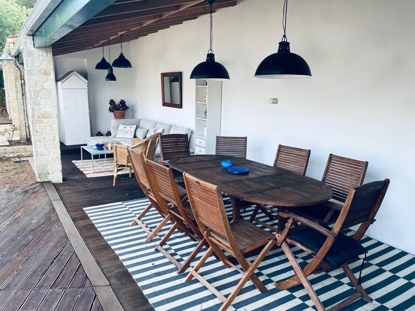 Un área de comedor al aire libre con mesa y sillas de madera, alfombra a rayas, lámparas colgantes y una pared de piedra.