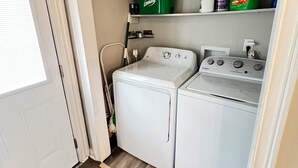 Een wasruimte met een voorlader wasmachine en droger, een bezem, een dweil en schoonmaakspullen op een plank.