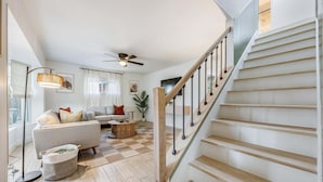 Un salón amplio con escaleras, un ventilador de techo y un ventanal con cortinas.