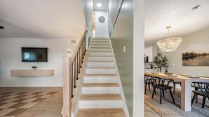 Una sala de estar moderna con una escalera, un televisor de pantalla plana, un comedor con una araña y un cuadro en la pared.