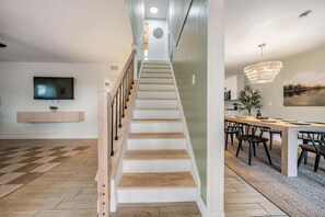 Ein modernes Wohnzimmer mit Treppe, einem Flachbildfernseher, einem Essbereich mit einer Chandelüster und einem Gemälde an der Wand.