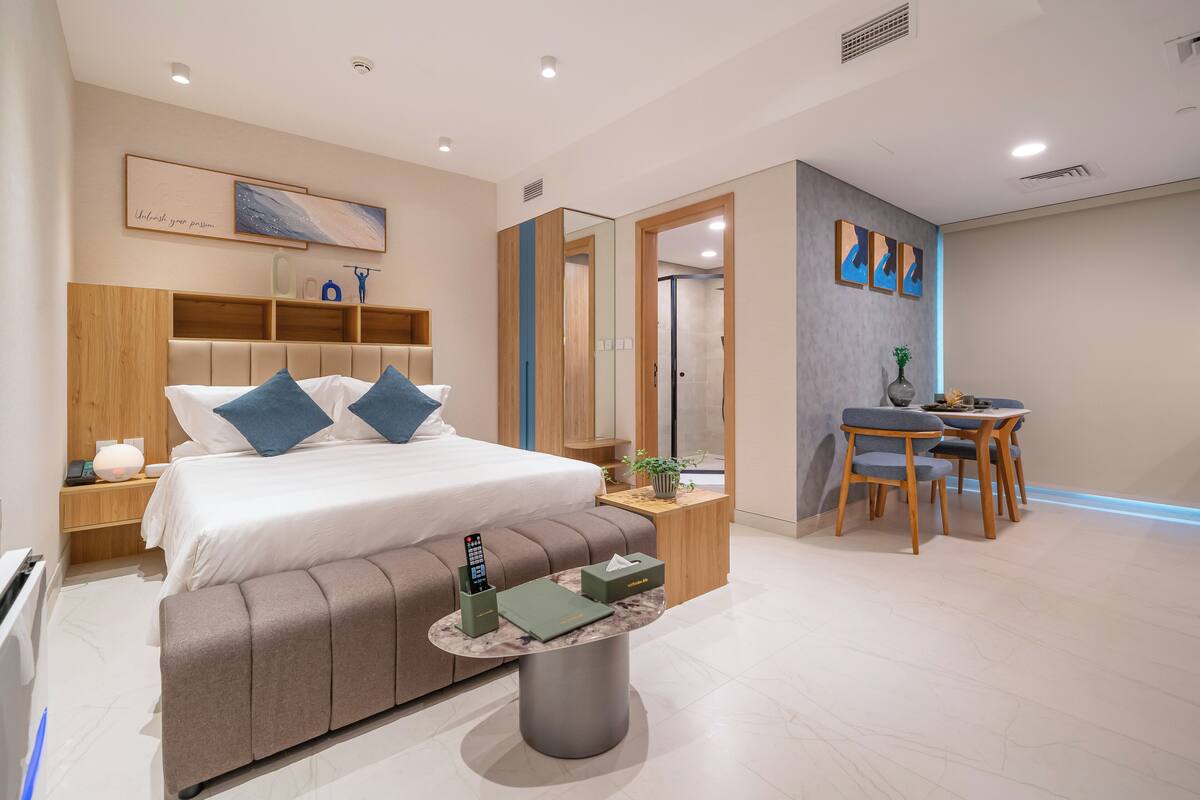 A modern hotel room with a large bed, bedside tables, a desk with chairs, and a shower area.