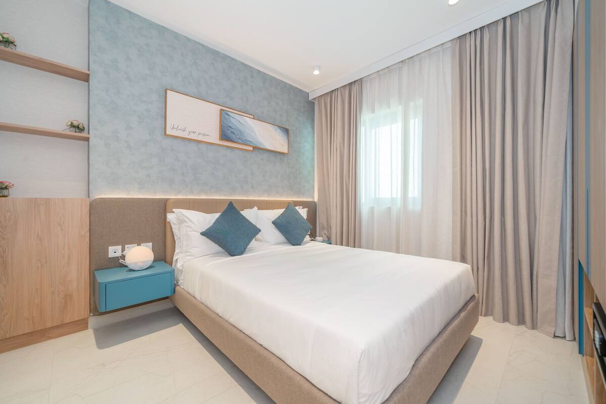 A modern bedroom with a large bed, a nightstand, a wall-mounted shelf, and a window with curtains.