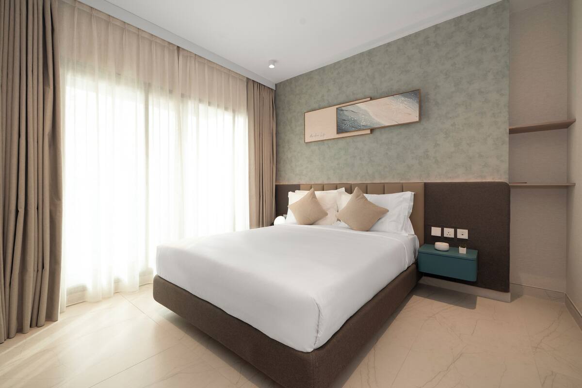 A modern hotel room with a large bed, a nightstand, a wall-mounted artwork, and a view of the outdoors through sheer curtains.
