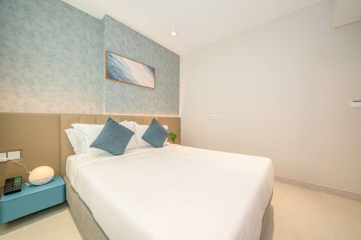 A modern hotel room with a neatly made bed, a bedside lamp, a phone on a nightstand, and a wall-mounted painting.