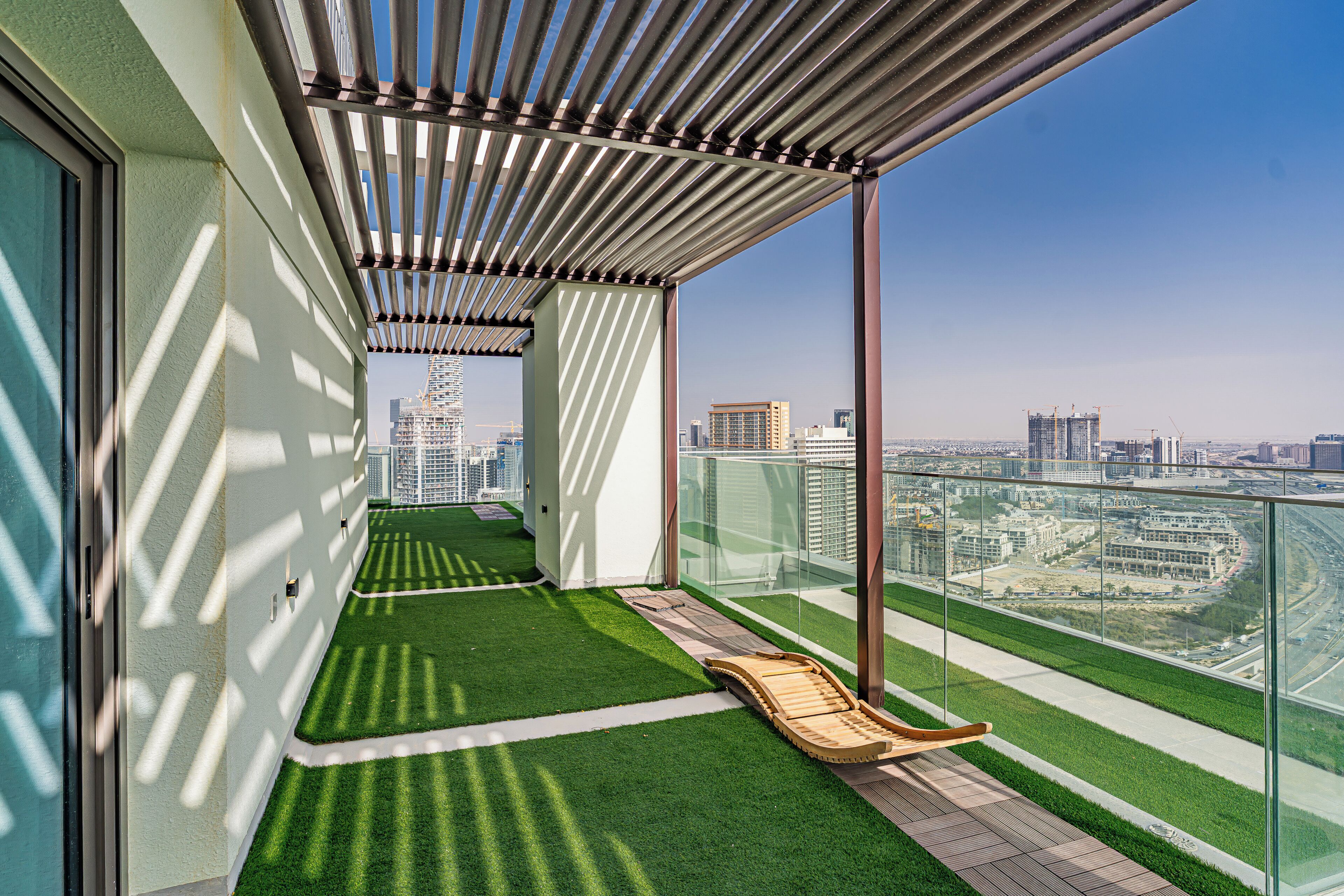 A balcony with a view of a cityscape, a grassy area, and a wooden lounge chair.