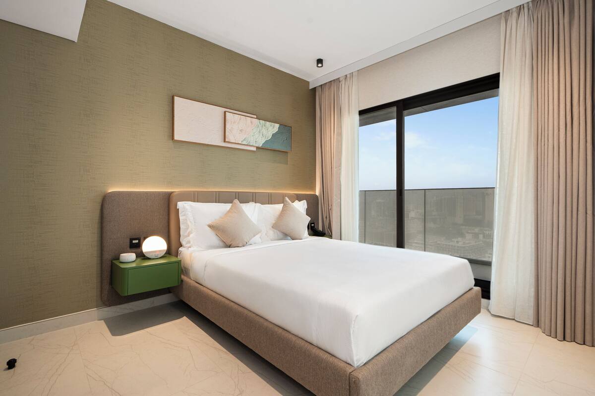 A modern hotel room with a large bed, a nightstand with a lamp, a cityscape view through the window, and a green accent wall.