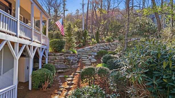 A house with a stone pathway leading to a wooden deck, surrounded by dense foliage and a flag. - Forget-Me-Not: Spacious 6-Bedroom Retreat, Walk to Downtown (Highlands)