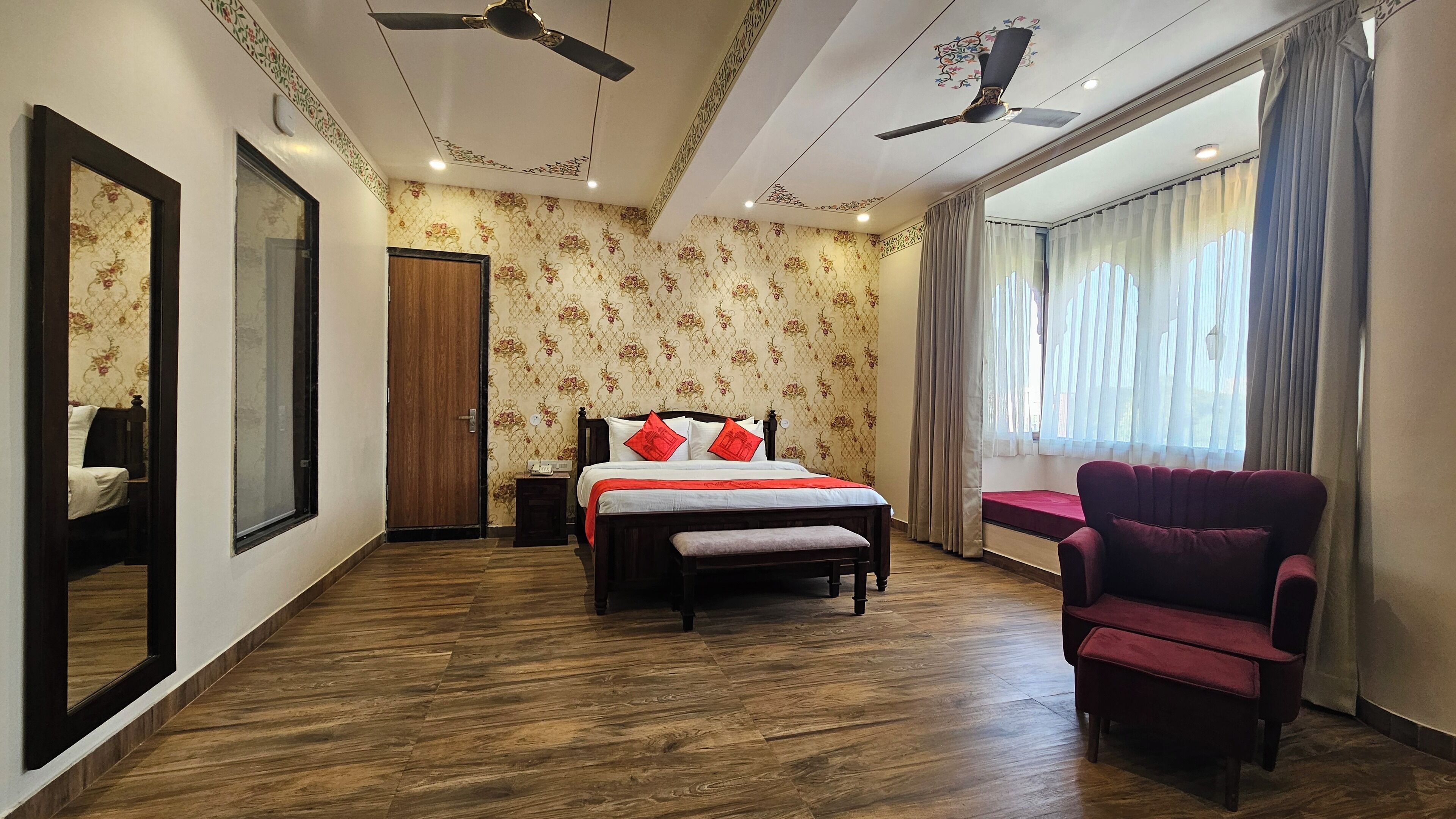 A bedroom with a wooden floor, a bed with red pillows, a chair, a ceiling fan, and a window with curtains.