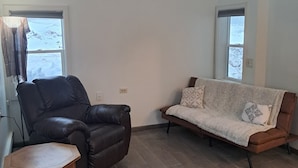 Ein Wohnzimmer mit einem braunen Ledersessel, einem Holztisch und einem braunen Sofa mit gemusterten Kissen. Zwei Fenster mit Vorhängen und ein Holzboden.
