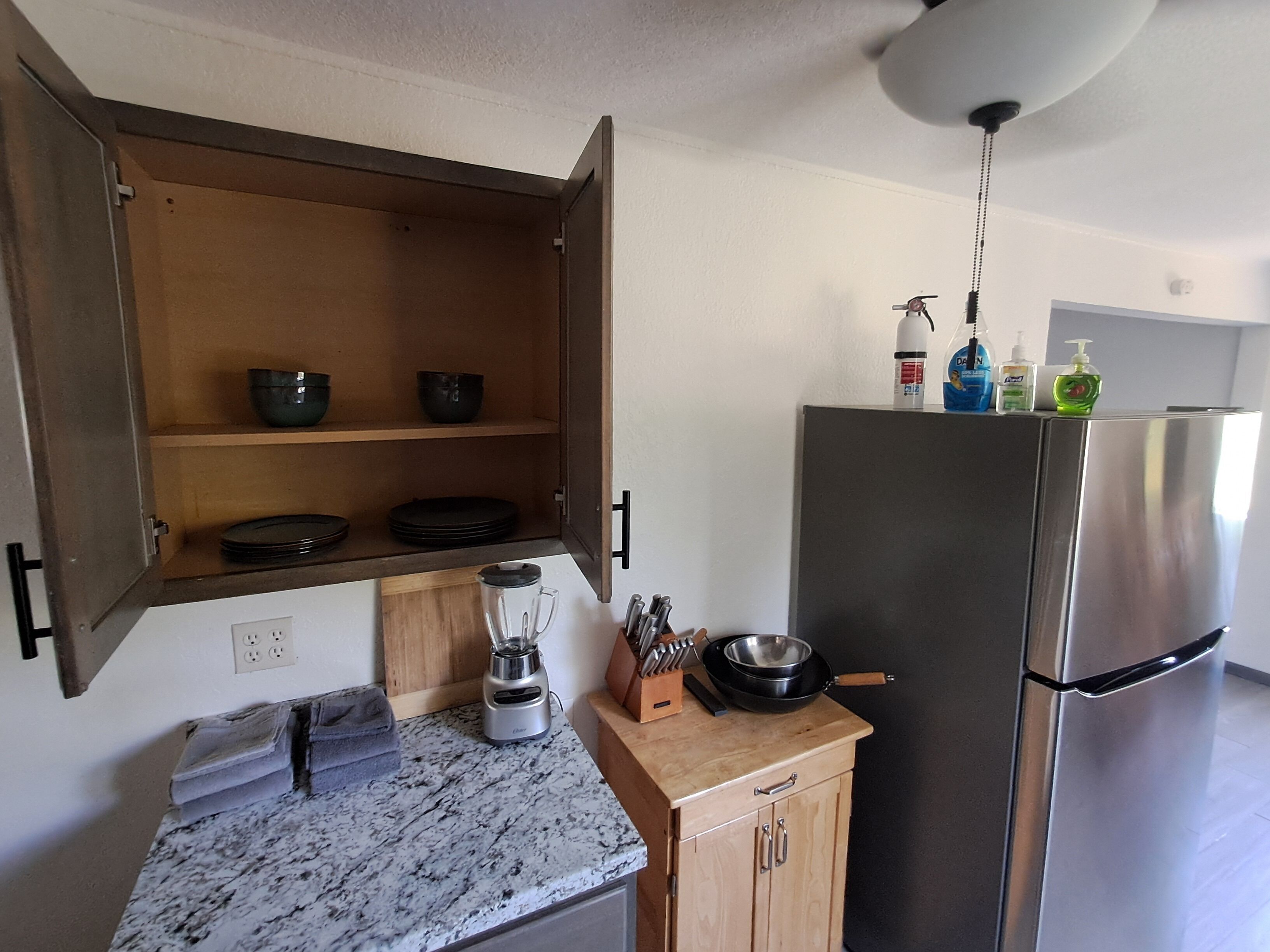 A kitchen with a marble countertop, a wooden cabinet with open shelves, a stainless steel refrigerator, and a ceiling light fixture.