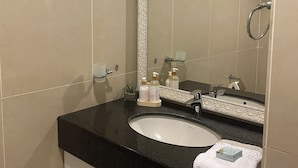 Un baño con encimera negra, lavamanos blanco y un espejo grande. Hay artículos de aseo en la encimera y una toalla en el lavamanos.