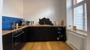Een kleine keuken met een houten aanrecht, zwarte kastjes, een inbouwoven en een raam met zonwering.