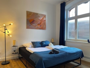 Une chambre à coucher avec un lit, une lampe sur pied, un tableau et une fenêtre avec des rideaux.