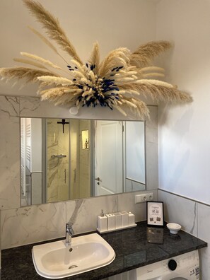 Un baño con tocador de mármol, lavamanos, espejo y un arreglo de plantas decorativas sobre el espejo.