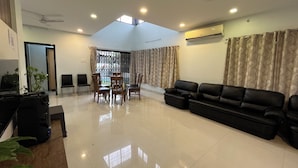 A spacious living area with a black leather sofa, a dining table with chairs, a TV, and an air conditioning unit. - Aarna Villa nr Valvan Dam (Lonavala)