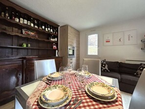 Comedor con mantel a cuadros, platos, vasos y cubiertos. Una estantería de madera con botellas se encuentra a la izquierda.