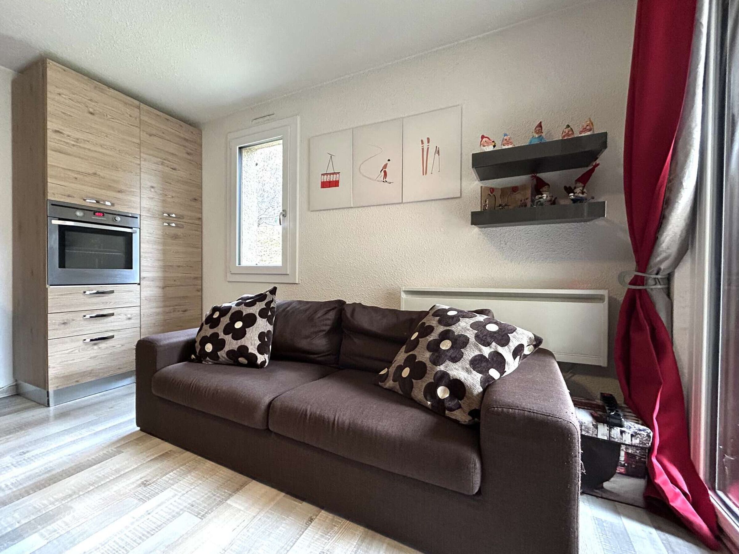 A modern living room with a brown sofa, two patterned pillows, a built-in oven, and a window with a view of the outside.