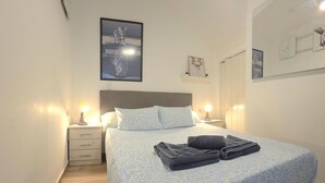 Ein ordentlich bezogenes Bett mit einem gemusterten Überwurf, zwei Nachttischlampen, ein Nachttisch mit Schublade und ein Poster an der Wand.