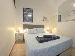 Ein ordentlich bezogenes Bett mit einem gemusterten Überwurf, zwei Nachttischlampen, ein Nachttisch mit Schublade und ein Poster an der Wand.