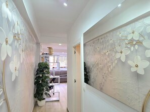 Ein Flur mit einer weißen Wand und einem großen Blumenbild. Im Hintergrund steht ein Blumentopf und eine weiße Bank.