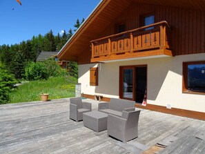 Eine Holzhütte mit Balkon, Sitzgelegenheit im Freien und Blick auf einen bewaldeten Bereich.