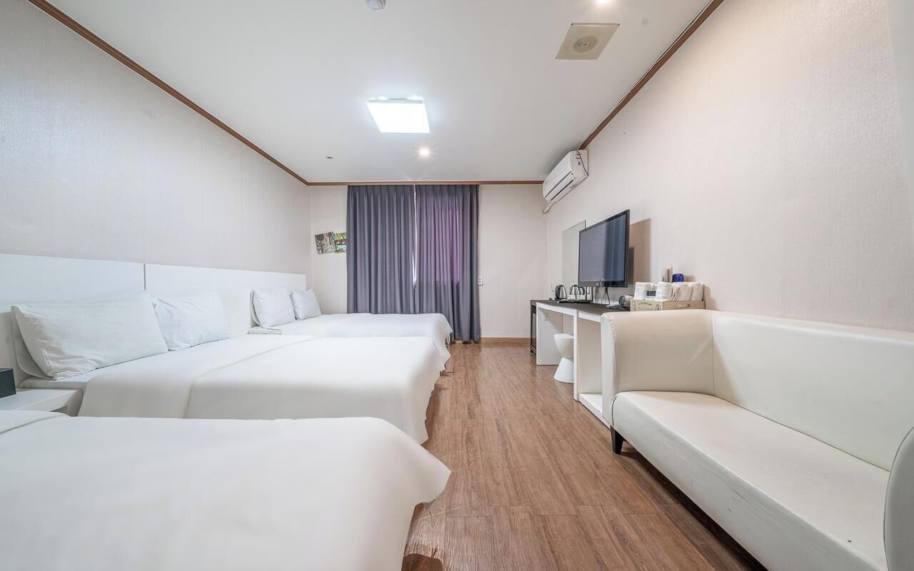 Habitación de hotel con dos camas, un sofá blanco, un escritorio con televisor y un equipo de aire acondicionado.
