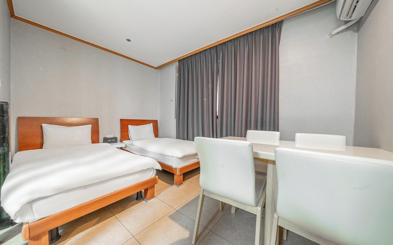 A hotel room with two single beds, a small desk with a chair, and a large window with curtains.
