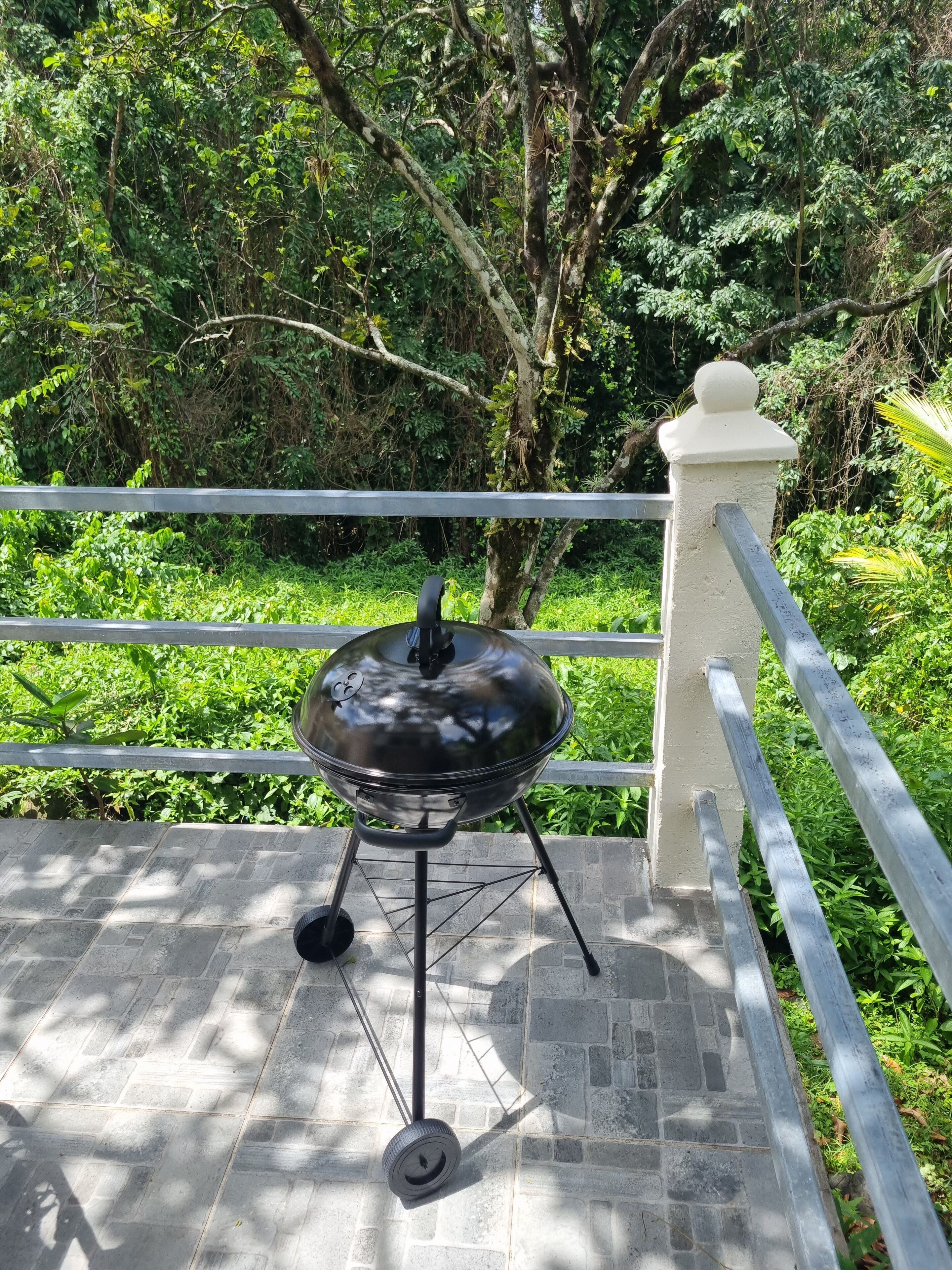 A barbecue grill on a tiled patio with a metal railing and lush greenery in the background.